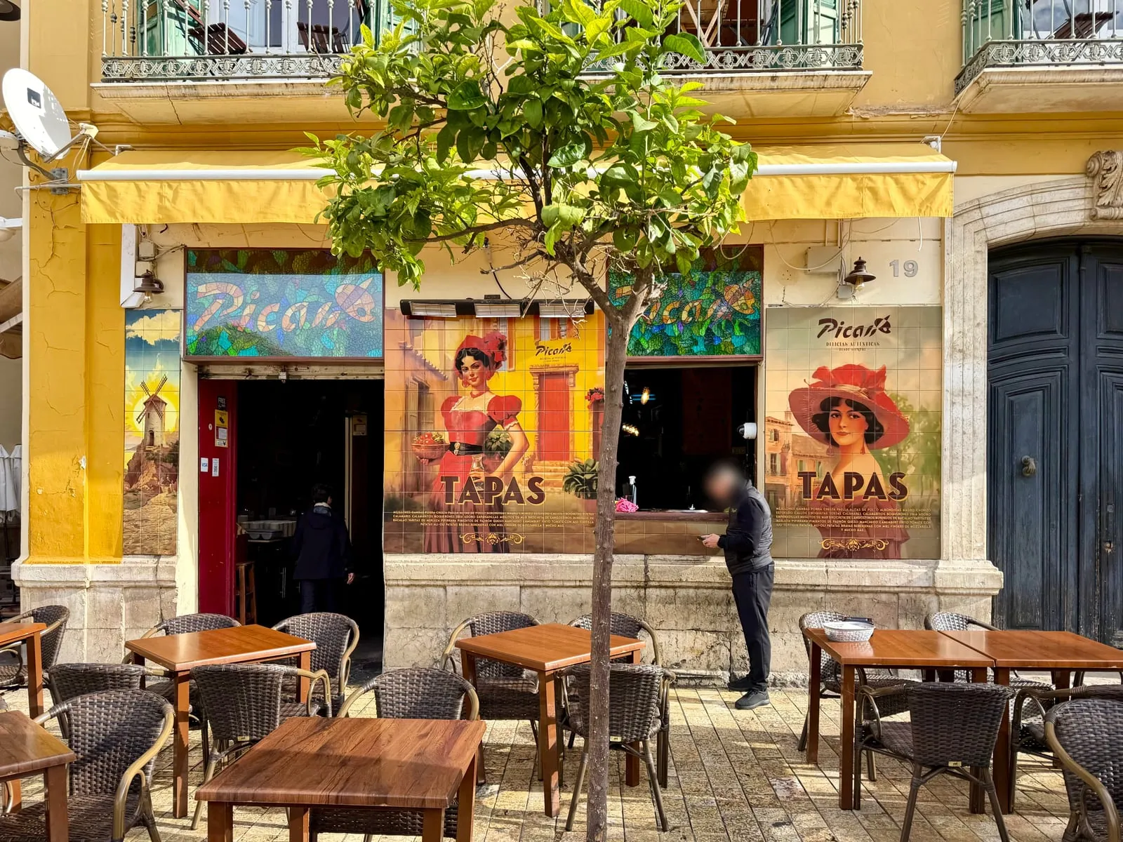 The front of Picas Brunch & Tapas at Plaza de la Merced 19: stained-glass Picas signs, tile posters above the word TAPAS, an orange tree and terrace tables
